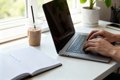 Mains sur le clavier d'un ordinateur portable, à côté d'un carnet ouvert et d'un café, près d'une fenêtre.