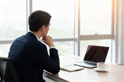 Personne pensive de dos à un bureau près d'une fenêtre, avec ordinateur portable, carnet ouvert et tasse de café