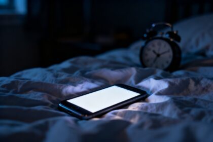 Smartphone allumé posé sur un lit défait dans une chambre d'adolescent plongée dans l'obscurité.