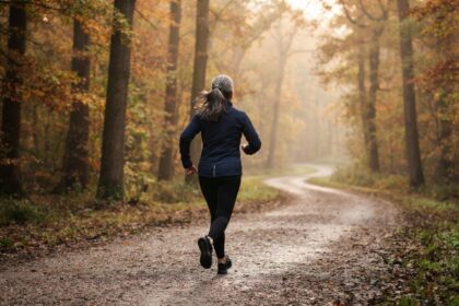 Une femme aux cheveux gris, vue de dos, court sur un chemin forestier brumeux aux couleurs d'automne.