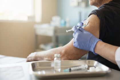 Un soignant ganté vaccine le bras d'une personne âgée, flacon et seringue posés sur un plateau médical