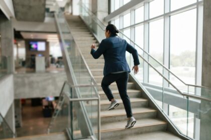 Une personne en tenue de bureau monte un escalier vitré d'un pas rapide, baskets aux pieds, en pleine lumière du jour.