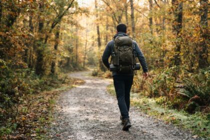Un homme vu de dos marche sur un sentier forestier en automne, un sac à dos chargé sur le dos.