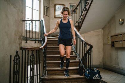 Femme en tenue de sport descendant un escalier d'immeuble en grimaçant, rouleau de massage et sac de sport au pied des marches